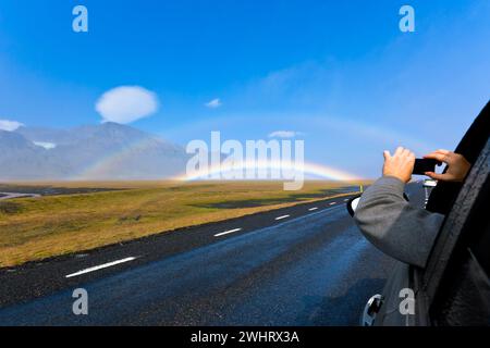 Mann in einem Auto schießt isländische Landschaft mit doppeltem Regenbogen Stockfoto