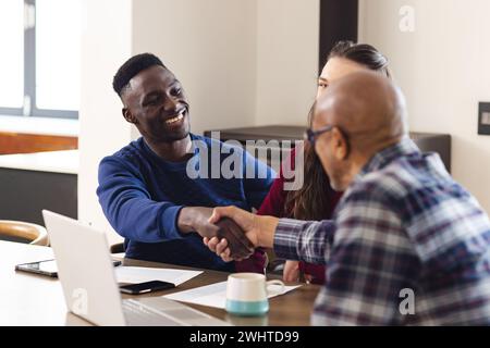 Diverse Paar- und birassische männliche Finanzberater, die Laptop benutzen und zu Hause die Hände schütteln Stockfoto