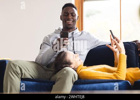 Glückliches, vielseitiges Paar, das zu Hause mit Smartphones auf dem Sofa sitzt Stockfoto