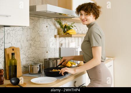 Lächelnde Frau legt gekochte Pasta mit Pilzsauce aus einer Pfanne auf einen Teller Stockfoto