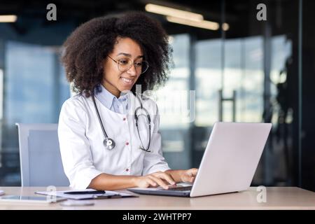 Selbstbewusste afroamerikanische Ärztin in medizinischer Uniform mit Laptop am Arbeitsplatz. Lächelnder Therapeut mit Stethoskop, der Medikamente nach Internetkonsultation des Kunden in der Klinik verordnet. Stockfoto