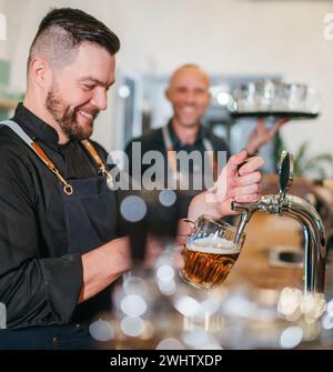 Stilvoller bärtiger Barkeeper in schwarzer Uniform, der Bier an der Bar anzapft und der Kellner mit Tablett plauderte miteinander und lächelte miteinander. Erfolgreiches People Team Wo Stockfoto