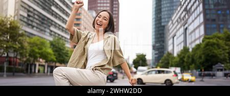 Porträt einer glücklichen asiatischen Frau, tanzen und Freude fühlen, triumphieren, die Hand in Siegesgeste hochheben, auf der Straße feiern Stockfoto