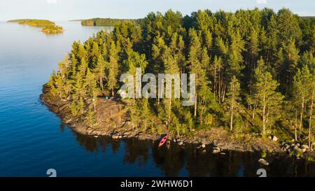 Drohnenaufnahme eines friedlichen Campingplatzes, eingebettet zwischen hohen Kiefern an einem felsigen Seeufer, mit einem Kajak am Wasser im sanften Abendlicht. Stockfoto