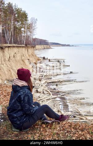 Frau in warmen Kleidern, sitzt auf einer Klippe über der Küste, bewundert die Natur, genießt die Einsamkeit. Aktiver Lifestyle-Moment. Das Konzept, bei jedem Wetter zu reisen Stockfoto