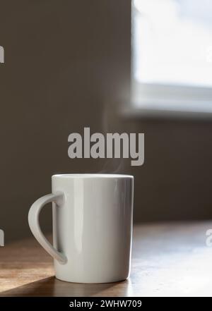 Ein einsamer weißer Kaffeetasse sitzt auf einem Holztisch am Fenster des Apartments im Morgenlicht. Dampf, der aus einer Morgenkaffee-Tasse kommt, die Konc Stockfoto