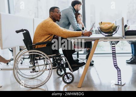 Hübscher schwarzer, männlicher Mitarbeiter sitzt im Rollstuhl und sieht sich etwas an, während seine Kollegen an einem neuen Projekt am Schreibtisch kooperieren. Stockfoto