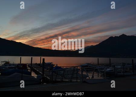 Panoramablick auf den roten Sonnenuntergang über See und Berge. Dunkle Holzpier mit verankerten Motorbooten, Sonnenstrahlen reflektiert auf dem Wasser, gestreifte Wolken am Himmel. Stockfoto