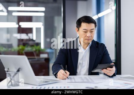 Ernsthaft denkender Geschäftsmann Buchhalter, der Finanzberichte überprüft, reifer konzentrierter asiatischer Mann hinter Papierarbeit, Taschenrechner in Händen, Arbeiter im Business-Anzug mit Laptop im Büro. Stockfoto