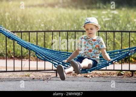 Der träumerische kleine Junge schwingt im Sommer in einer Hängematte im Park. Das Konzept der Kindererholung und -Freizeit in der Stadt. Stockfoto