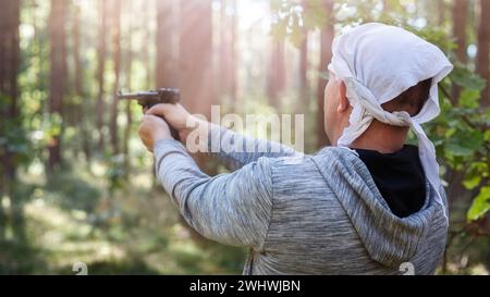 Ein Mann schießt eine Pistole im Wald zwischen den Bäumen, die von Sonnenlicht beleuchtet werden. Rückansicht, unscharfer Hintergrund. Stockfoto