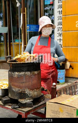 Eine Straßenverkäuferin, die frittierte Kartoffeln verkauft, erwartet einen hungrigen Kunden vor dem Sera Kloster in Lhasa. Stockfoto