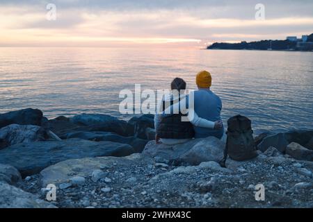 Mann und Frau sitzen auf riesigen Steinen am Wasser und halten abends Hände. Stockfoto