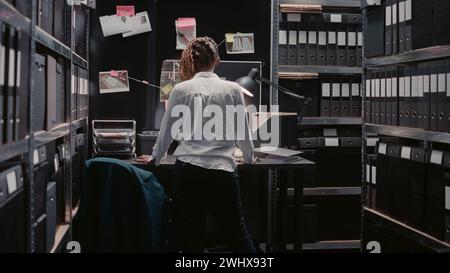 Anwaltsbeamter liest Strafregister im Archiv, überprüft verdächtige Fotos und findet Hinweise. Weiblicher Ermittler, der Ordner mit Verbrechen hält, verdächtige Hintergrundüberprüfung durchführt. Stockfoto