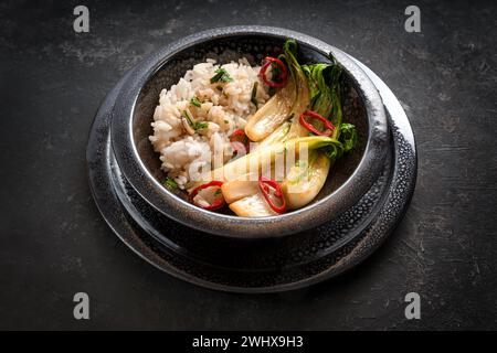 Pak Choi (Chinakohl) mit Reis in einer schwarzen Keramikschale mit Knoblauch, Ingwer, rotem Chili und Kräutern auf dunklem Hintergrund Stockfoto