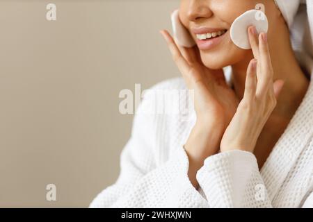 Eine hübsche Frau im Gewand und Handtuch auf dem Kopf wischt sich die Haut ihres Gesichts mit Wattepads ab. Reinigung und gesunde Körperpflege nach SH Stockfoto