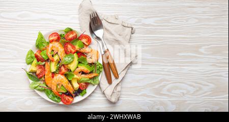 Gesunder Salat mit gegrillten Garnelen, Avocado, Kirschtomaten und grünen Blättern auf weißem Teller mit Besteck auf weißem Holz rustikal Stockfoto