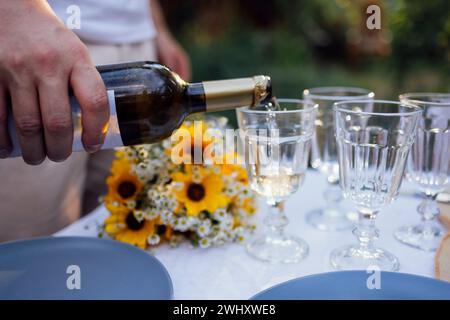 Der Mann gießt Weißwein aus der dunklen Flasche draußen im Garten in Gläser. Nahaufnahme der männlichen Hand und des Bechers. Stockfoto