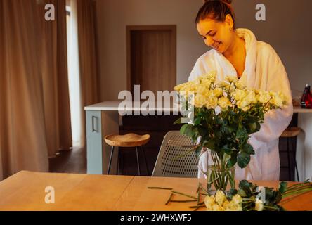 Die junge charmante Frau im weißen Bademantel schneidet weiße Rosen ab und legt sie in eine Glasvase. Stockfoto