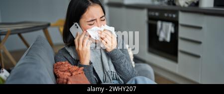Porträt einer kranken jungen koreanischen Frau, die sich krank fühlt, niest und Serviette hält, zu Hause krank bleibt, erkältet ist. Auf dem Mobiltelefon sprechen Stockfoto