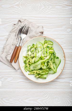 Gesunde vegane grüne Avocado Salatschüssel mit geschnittenen Gurken, Edamame Bohnen, Olivenöl und Kräutern auf Keramikplatte Draufsicht, weiß Stockfoto