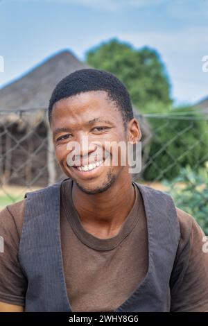 Der ehrenamtliche Sozialarbeiter des Dorfes, ein junger, lächelnder afrikanischer Mann mit einer Weste im Hof, der sich ausruhen lässt, traditionelle Hütte und Rondavel im Hintergrund Stockfoto