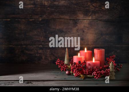 Dritter Adventskranz mit roten Kerzen, drei sind beleuchtet, Dekoration mit Beeren, Weihnachtskugeln und kleinen Holzbäumen, dunkle Rusti Stockfoto