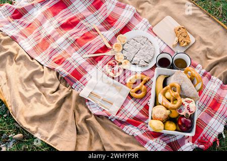 Ein Korb mit Gebäck und Obst steht neben Kaffeetassen auf einer Decke auf grünem Gras Stockfoto