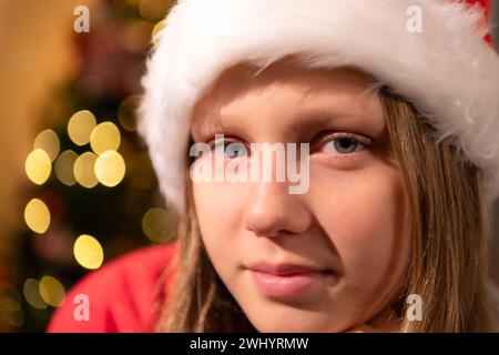 Porträt eines Teenagers im Weihnachtsmann-Hut, der auf einem Stuhl vor dem Weihnachtsbaum sitzt, frohe Weihnachten und frohe Feiertage! Stockfoto