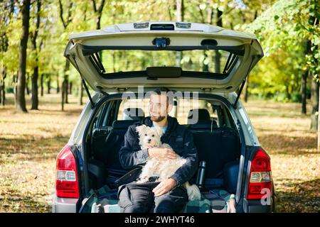 Junger Mann mit Hund, der sich entspannt und die Natur genießt, sitzt im Kofferraum Stockfoto