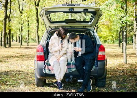 Liebe junge Familie, die seinen süßen Hund streichelt und küsst, während sie im Kofferraum sitzt und sich im Wald entspannt Stockfoto