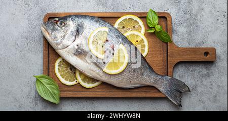 Roher, ungekochter Fisch dorado mit Zitrone und frischem Basilikum auf hölzernem Schneidebrett mit Messer auf rustikalem Steinhintergrund von oben Blick, kochen Stockfoto