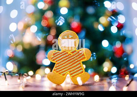 Lebkuchenmann in Schutzglasmaske auf dem Tisch im Licht mehrfarbiger Lichter am Tannenbaum. Unschärfe Stockfoto