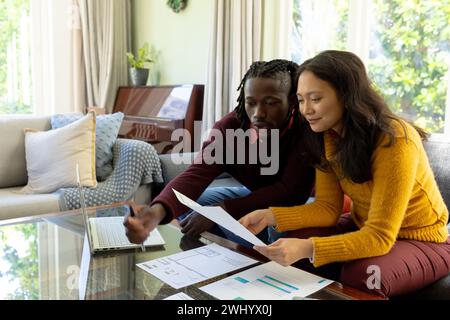 Glückliches, vielseitiges Paar, das auf der Couch sitzt und mit einem Laptop über Rechnungen und Finanzen im Wohnzimmer diskutiert Stockfoto