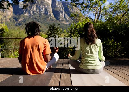 Rückansicht eines diversen Paares, das Yoga-Meditation praktiziert und auf Deck im sonnigen Garten sitzt Stockfoto