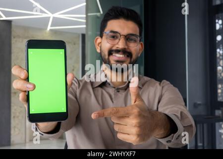 Glücklicher indischer Mann in Brille, der mit Zeigefinger auf den Handy-Bildschirm mit leerem Raum führt. Junger bärtiger Mann, der eine neue Anwendung empfiehlt und die Vorteile der Installation von Service genehmigt. Stockfoto