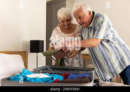 Glückliches kaukasisches Seniorenpaar, das Koffer packt und im sonnigen Schlafzimmer zu Hause lächelt Stockfoto