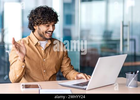 Freundlicher Programmierer aus dem Nahen Osten, der die Hand winkt, während er einen tragbaren Computer für die Online-Fernkommunikation verwendet. Positive professionelle Begrüßung Gesprächspartner vor Beginn des Geschäftsgesprächs. Stockfoto