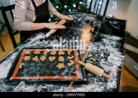 Koch rollt den Teig auf dem Tisch neben dem Tablett mit ausgeschnittenen Keksen aus. Abgeschnitten Stockfoto