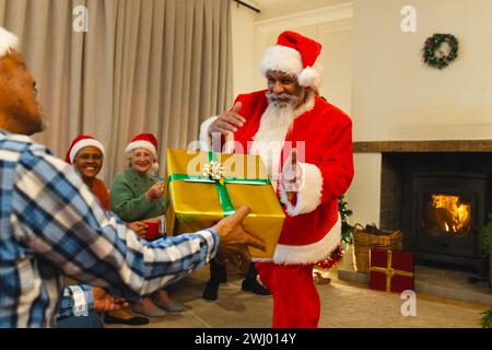 Glücklicher birassischer Mann in weihnachtsmannskleidung, der einem Freund neben dem Kamin zu Hause ein Geschenk schenkt Stockfoto