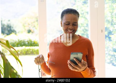 Glückliche afroamerikanische Seniorin mit Brille, mit Smartphone im sonnigen Wohnzimmer, Kopierraum Stockfoto
