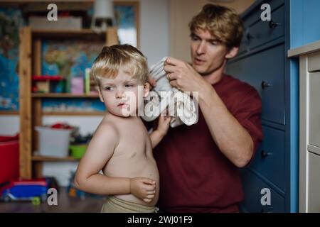 Vater zieht Hemd auf den kleinen Sohn und zieht sich morgens vom Schlafanzug. Der Junge ist traurig, weinend. Stockfoto