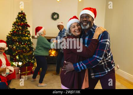 Zwei glückliche, vielseitige ältere Freunde in santa-Hüten, die sich im Wohnzimmer zu Hause umarmen Stockfoto