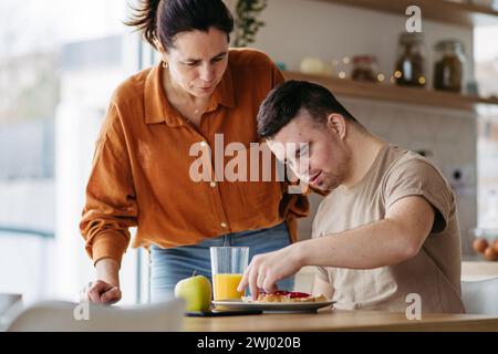 Junger Mann mit Down-Syndrom, der das Frühstück mit seiner Mutter zuhause zubereitet. Morgenroutine für Mann mit Down-Syndrom. Stockfoto