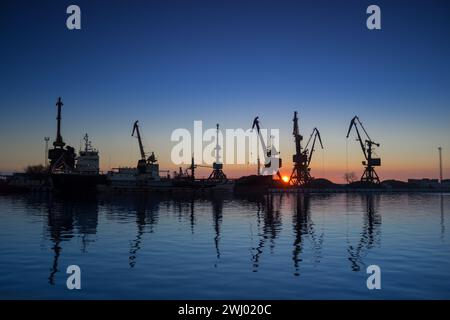 Sonnenaufgang am Hafen, Silhouetten von Kränen reflektieren im Wasser, Beginn des Frachtbetriebs, Lieferkettenlogistik, Import-Export-Industrie, Seefahrt Stockfoto