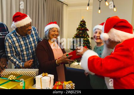 Glücklicher birassischer Mann in weihnachtsmannskleidung, der Freunden im Wohnzimmer zu Hause Geschenke schenkt Stockfoto