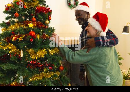 Zwei glückliche, vielseitige Seniorenfreunde in weihnachtsmützen, die den weihnachtsbaum zu Hause schmücken Stockfoto
