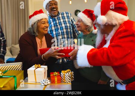 Glücklicher birassischer Mann in weihnachtsmannskleidung, der Freunden im Wohnzimmer zu Hause Geschenke schenkt Stockfoto