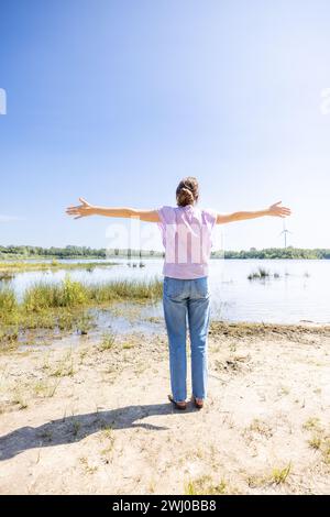 Dieses Bild zeigt eine Frau von hinten, während sie mit ausgestreckten Armen steht und mit Blick auf eine malerische Naturlandschaft steht. Sie scheint die Offenheit und Schönheit der Umgebung um sie herum zu begrüßen, zu der ein ruhiges Wasser, Grün und ein klarer blauer Himmel gehören. Die Präsenz von Windturbinen in der Ferne verleiht der ansonsten ruhigen und zeitlosen Szene einen Hauch von Modernität. Frau mit ausgestreckten Armen, die die Natur umarmen. Hochwertige Fotos Stockfoto