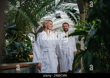 Wunderschönes Paar in Spa-Bademänteln, das im Gewächshaus des Hotels steht, sich ansieht und ein romantisches Wellness-Wochenende im Spa genießt. Valentinstag-Konzept Stockfoto